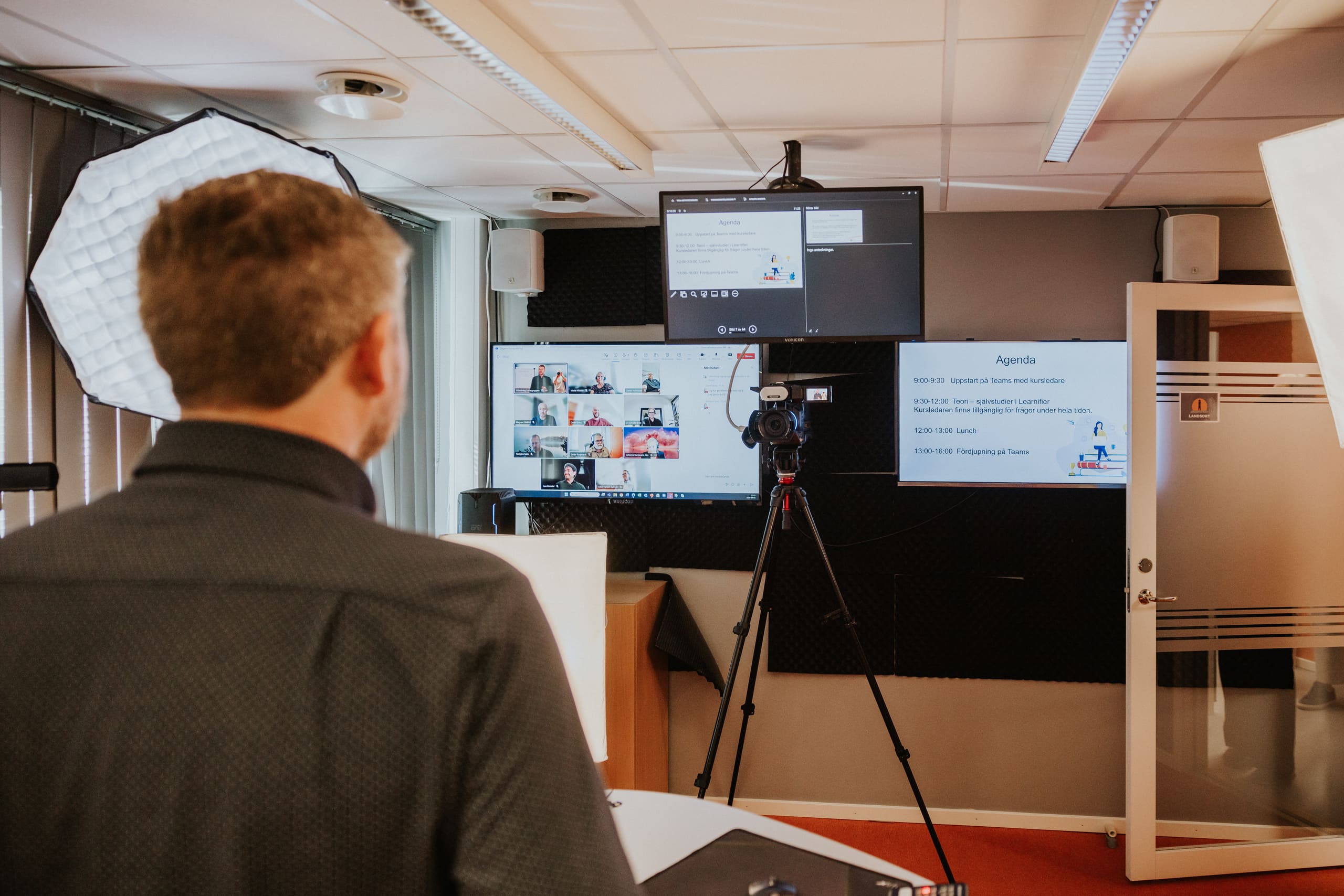 En kursledare håller en digital utbildning framför en kamera, med deltagare i ett videomöte och dagens agenda visad på flera skärmar.